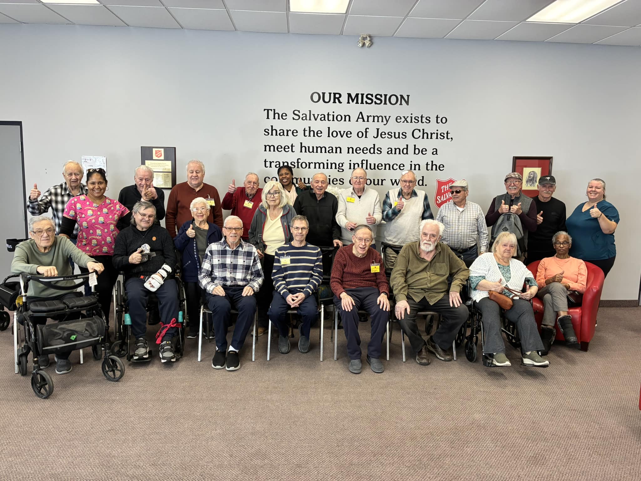 Group of program participants seated together in a community room, posed in front of a wall with The Salvation Army mission statement.